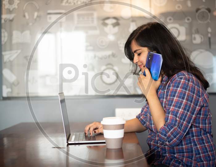 Portrait of a beautiful, young and intelligent-looking Indian Asian woman student wearing a shirt talks on phone as she works on her laptop