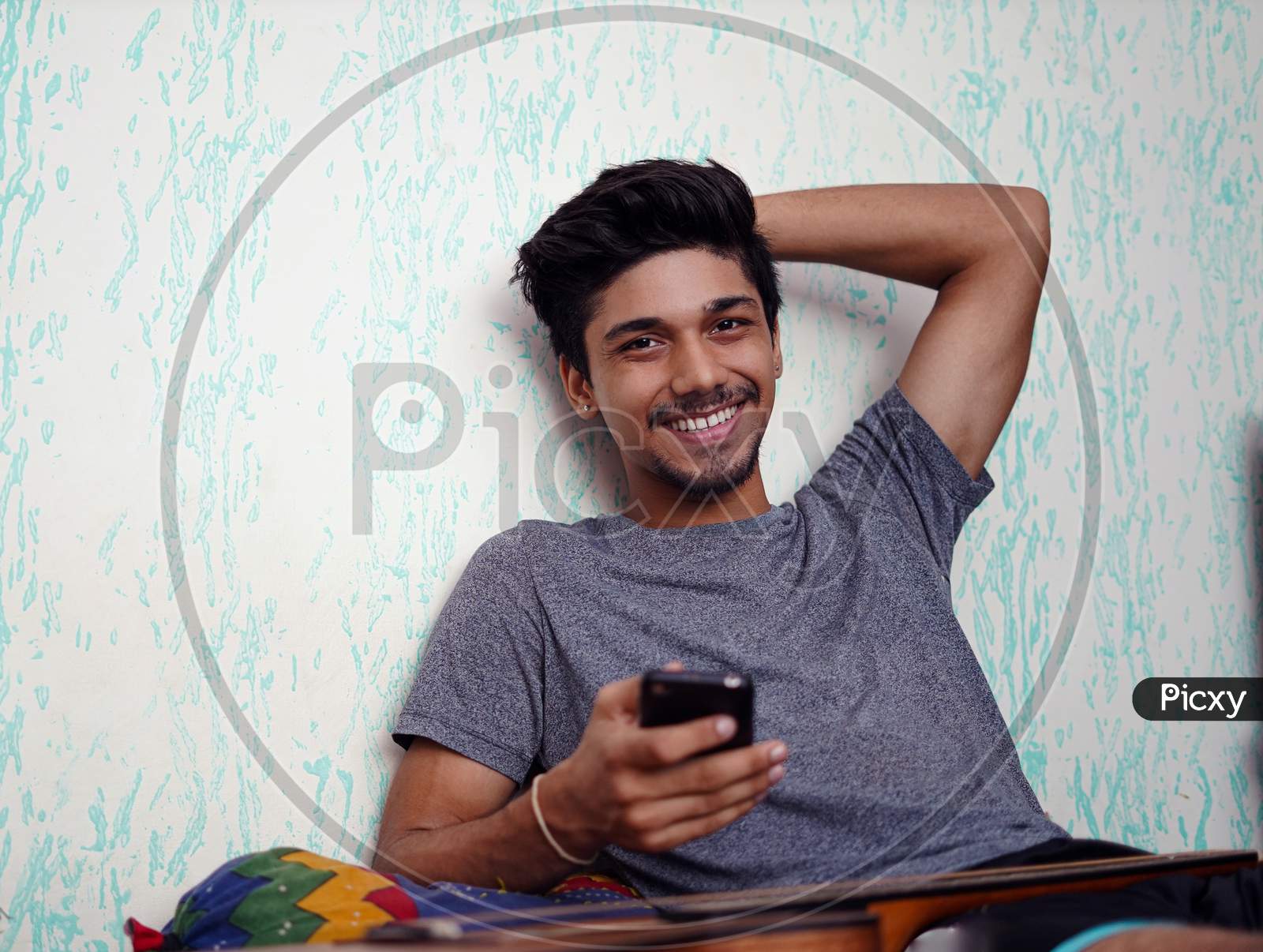 Young Indian Boy Smiling While Looking Into The Camera, With Phone In One Hand And The Other Behind His Head.