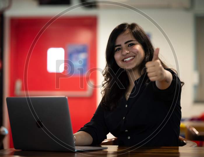 Portrait of a beautiful, young and intelligent-looking Indian Asian woman student gestures thumbs up wearing a shirt smiling as she works on her laptop at a workplace
