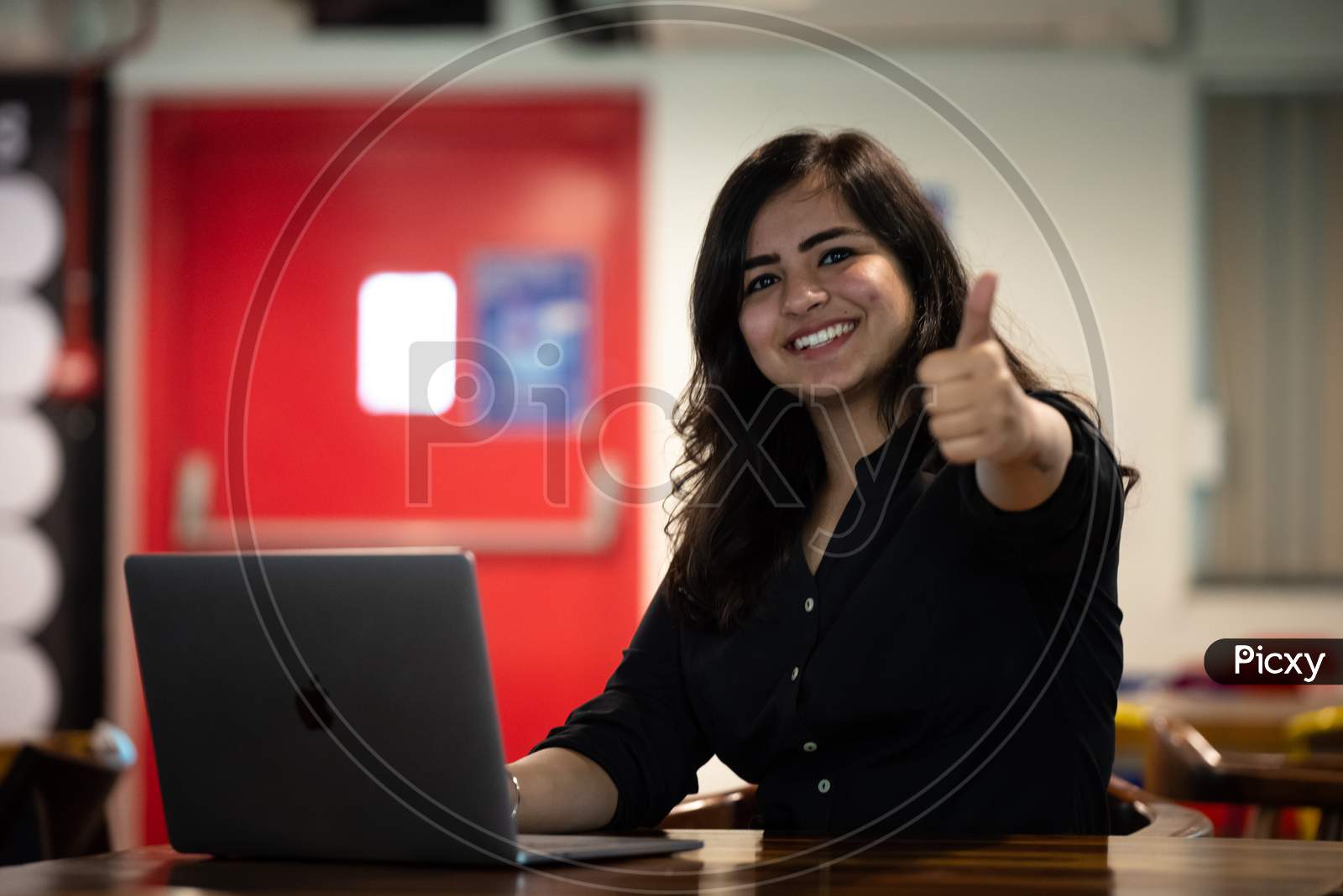 Portrait of a beautiful, young and intelligent-looking Indian Asian woman student gestures thumbs up wearing a shirt smiling as she works on her laptop at a workplace