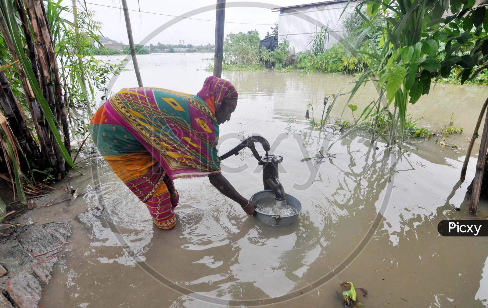 A Woman Collects Drinking Water From A Hand Pump With Her House Inundated With Flood Water In The Flood Affected Morigaon District Of Assam On June 25,2020.