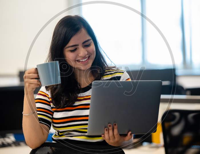 Portrait Of A Beautiful Young And Intelligent Looking Indian Asian Woman Student Wearing A T Shirt drinks coffee As She Works On Her Laptop