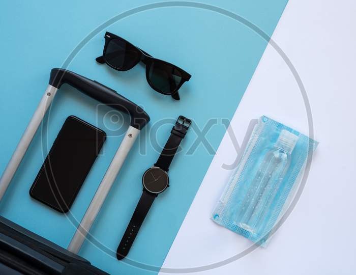 Accessories Of A Traveller On The Summer Holidays 2020 With The Coronavirus. Black Suitcase, Face Mask, Hydroalcoholic Gel, Clock, Smartphone And Sunglasses
