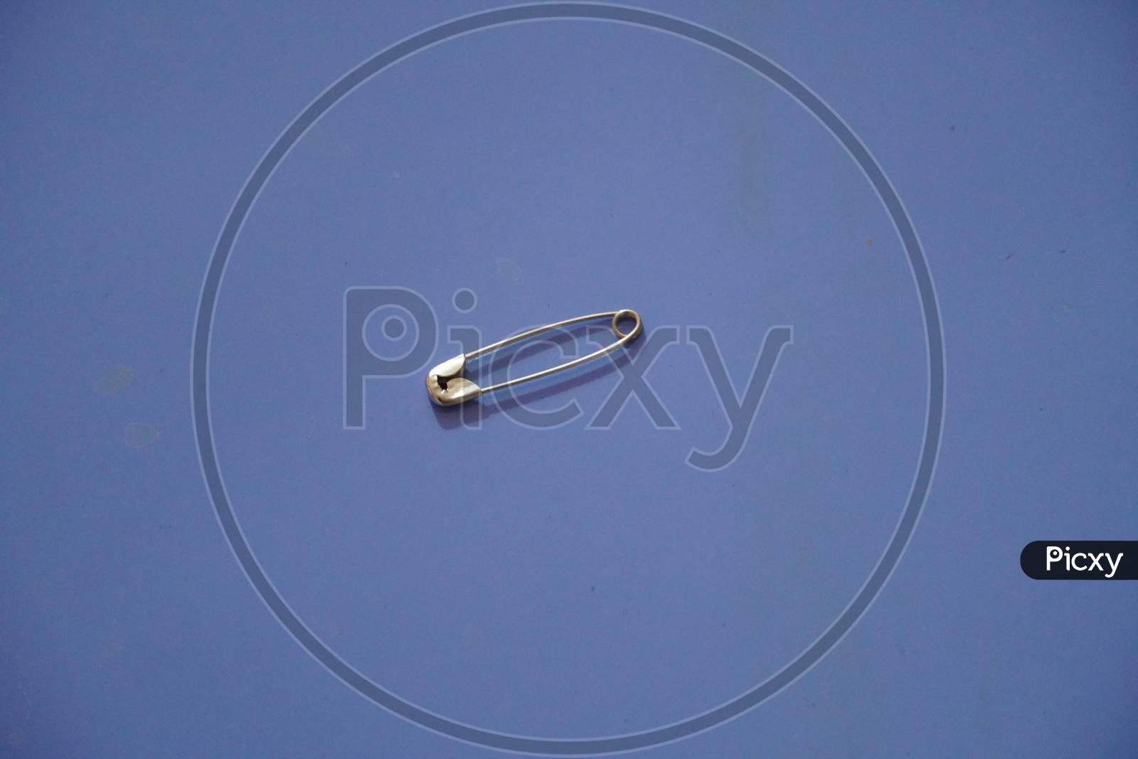 A Closed Silver Safety Pin Isolated On A Blue Background. Also Known As Shield Pin, Lingerie Pin, Clasp, Diaper Pin, Fastener And Sewing Pin.