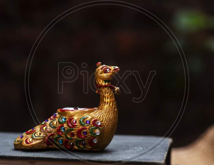 Gem studded beautiful and elegant showpiece of a golden Peacock sitting in Old Book with dark background. It is candle stand in golden color.