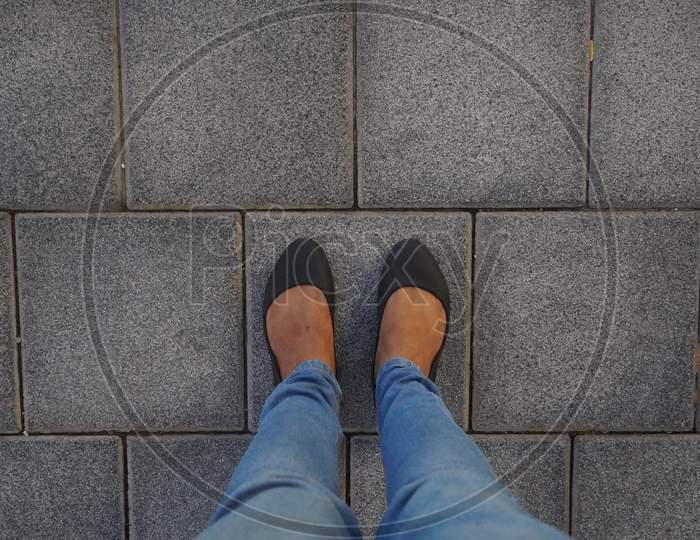 Women Black Casual Shoes Standing And Resting On Asphalt Concrete Floor With Square Tiles. Top View. Concrete Floor Texture Pattern Pavement Background. Selfie Female Of Feet And Legs Seen From Above.