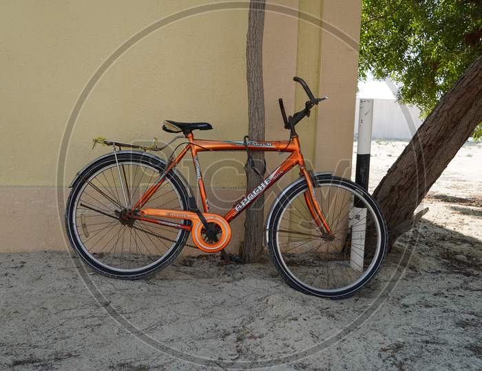 An Old Orange Bicycle Is Standing On The Sand Path Locked To A Tree. Old Apache Orange Cycle Chained To A Tree. Man'S Cycle With Lock In Tree In A Sandy Place. - Dubai Uae December 2019