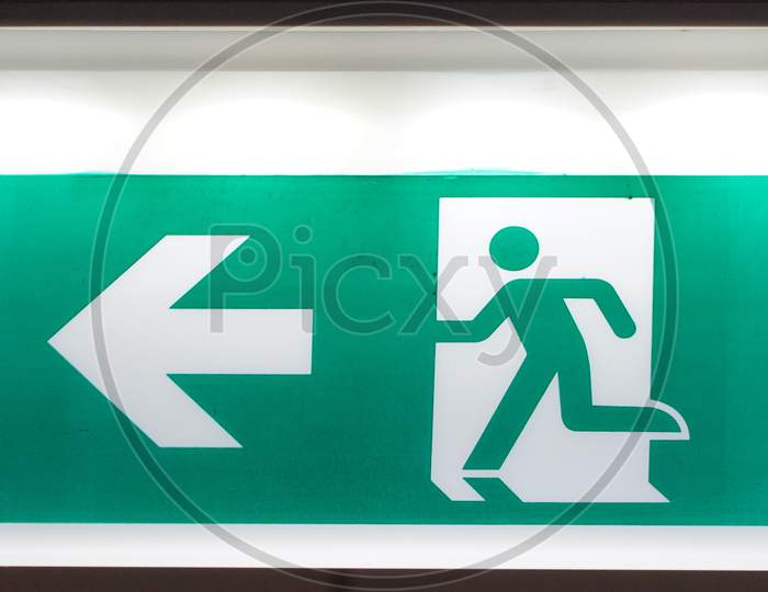 Glowing Emergency Exit Sign With Left Arrow At A Building. Safety First Concept. Copy Space Wallpaper. Exit Signs With Light In A Parking Of Building.