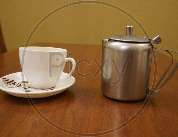 Old Dirty Used Aluminum Tea Pot Kettle With Dirty Used White Tea Mug. Antique Aluminum Kettle With Tea Cup And Empty Used Sugar Sachets. Dirty Coffee And Tea Cup Half Empty On Saucer On Table.