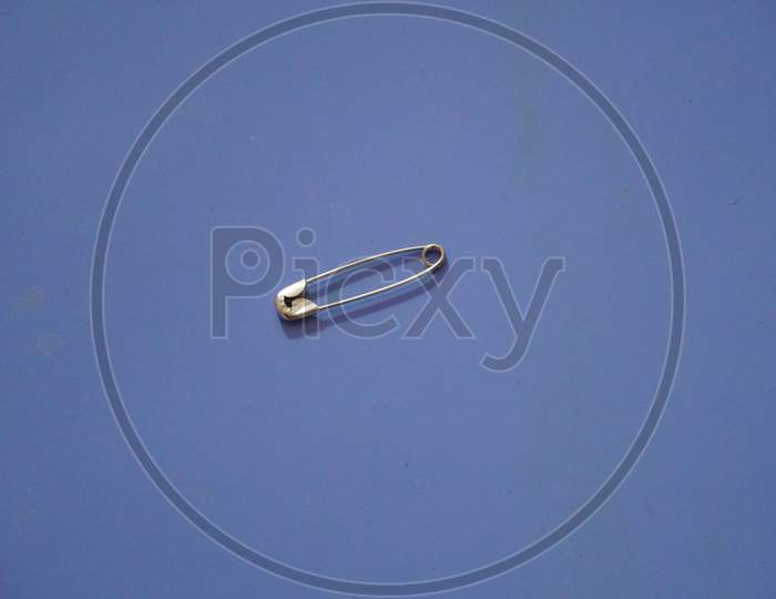 A Closed Silver Safety Pin Isolated On A Blue Background. Also Known As Shield Pin, Lingerie Pin, Clasp, Diaper Pin, Fastener And Sewing Pin.