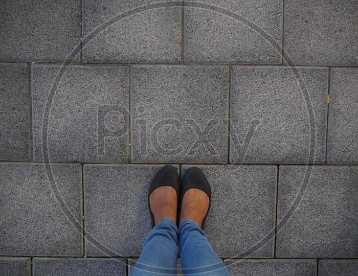 Women Black Casual Shoes Standing And Resting On Asphalt Concrete Floor With Square Tiles. Top View. Concrete Floor Texture Pattern Pavement Background. Selfie Female Of Feet And Legs Seen From Above.