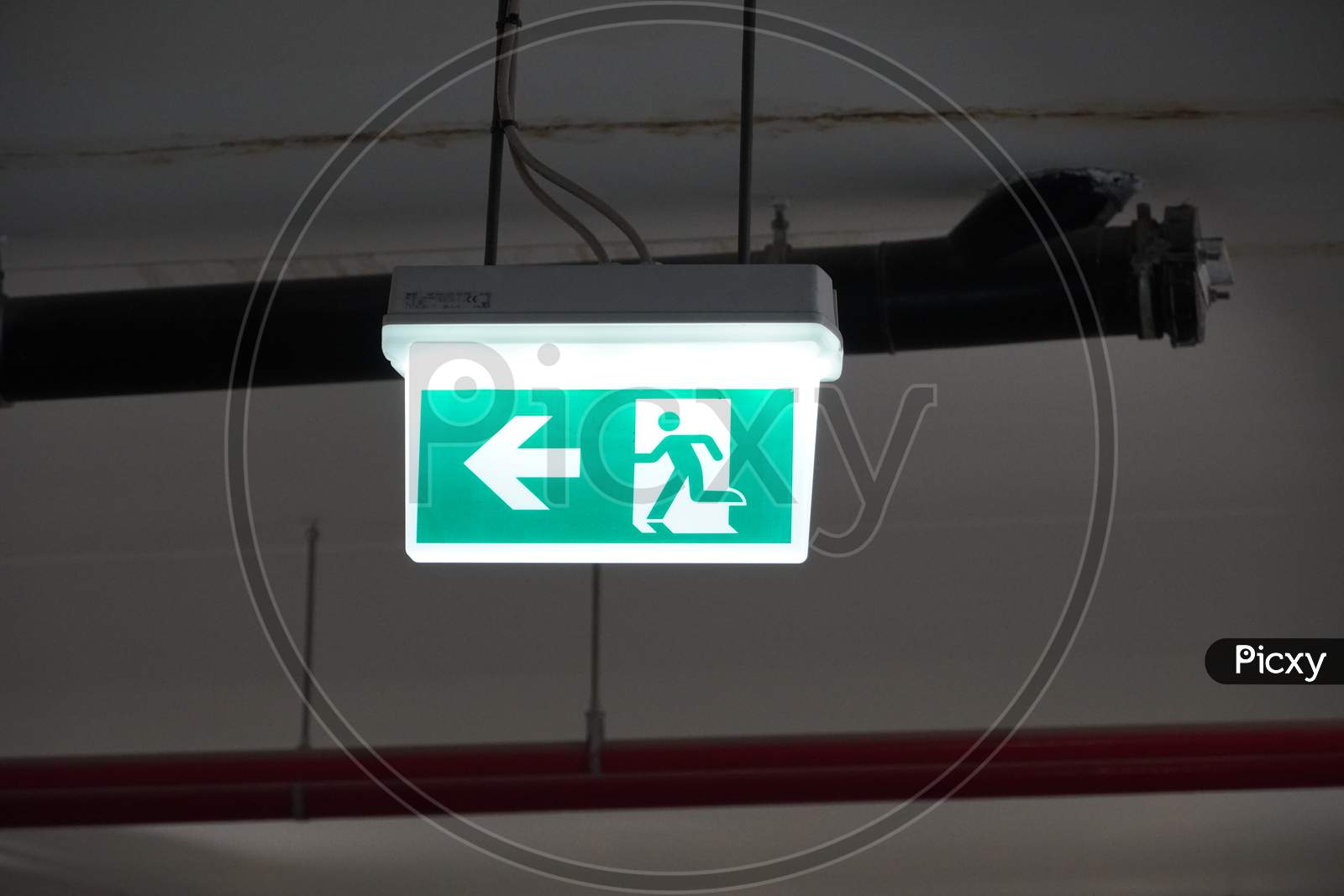Glowing Emergency Exit Sign With Left Arrow At A Building. Safety First Concept. Copy Space Wallpaper. Exit Signs With Light In A Parking Of Building.