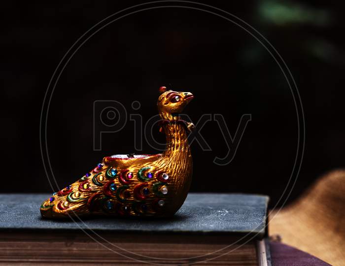 Gem studded beautiful and elegant showpiece of a golden Peacock sitting in Old Book with dark background. It is candle stand in golden color.