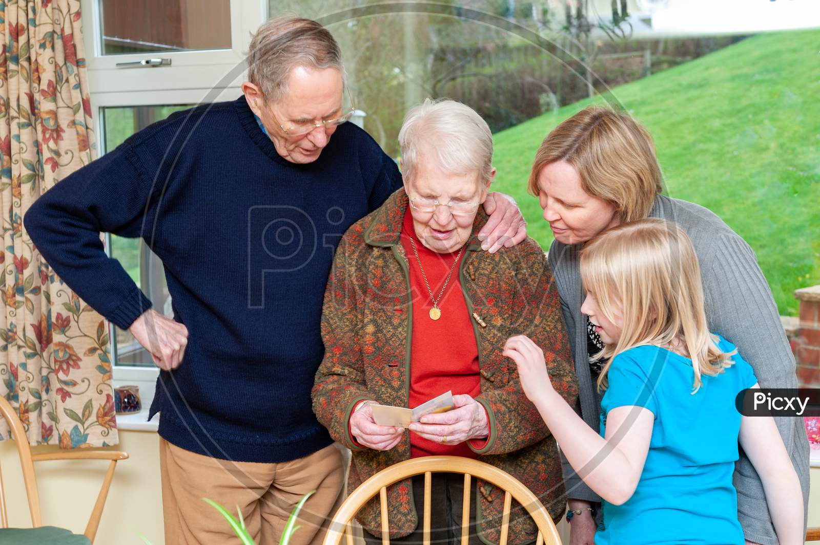 Three Generations Of A Family Gather Around While An Elderly Lady Reads A Birthday Card