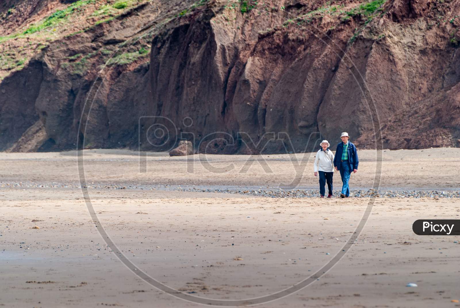 An Elderly Couple Walk Across An Empty Beach At The Base Of Tall Cliffs While Holding Hands