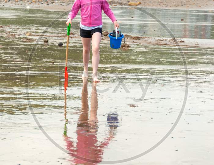 A Young Girl Wears A Pink Floppy Hat And Carries A Bucket And Spade Across A British Beach