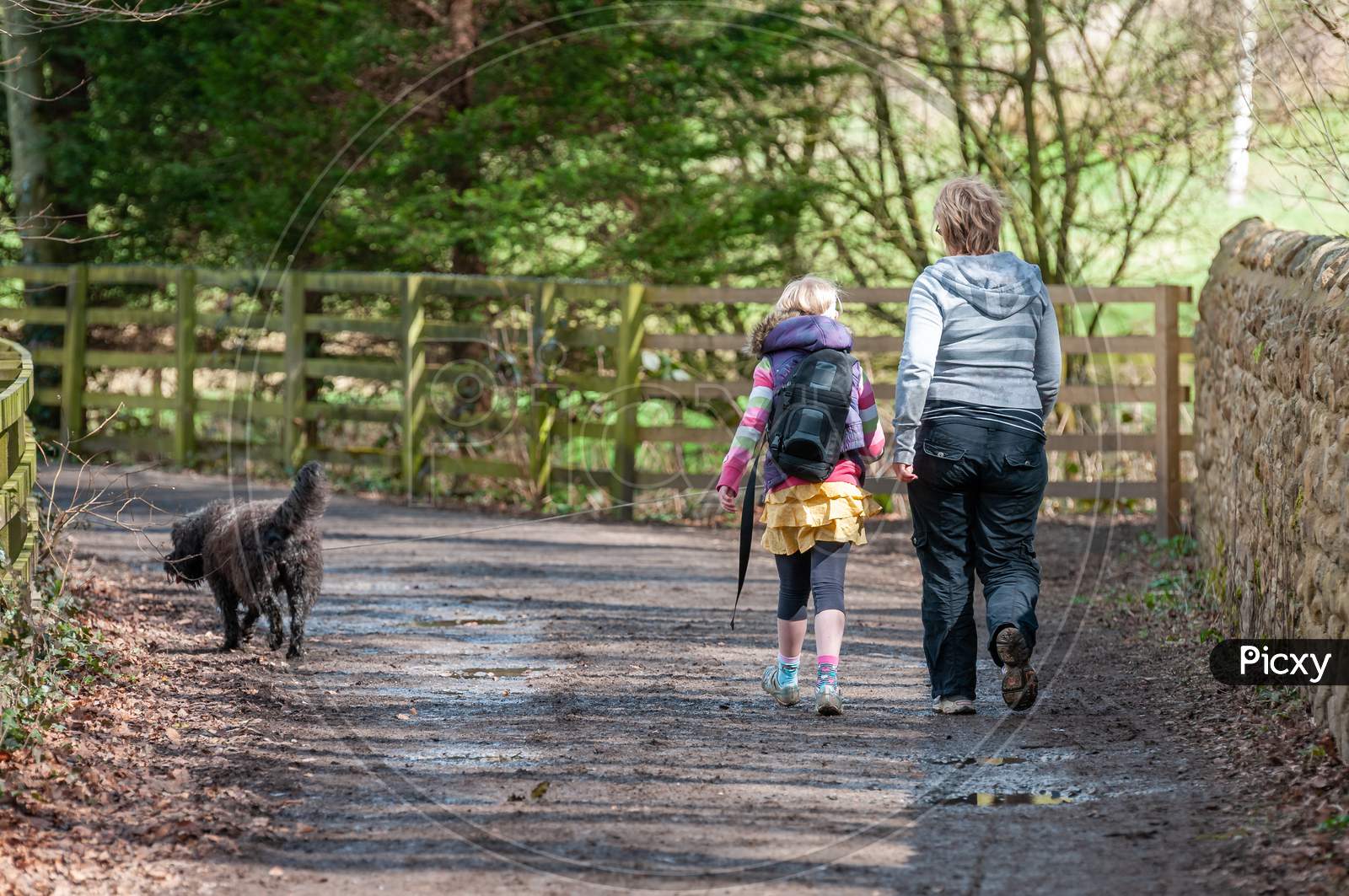 A Mother And Daughter Walk A Black Dog Along A Country Track