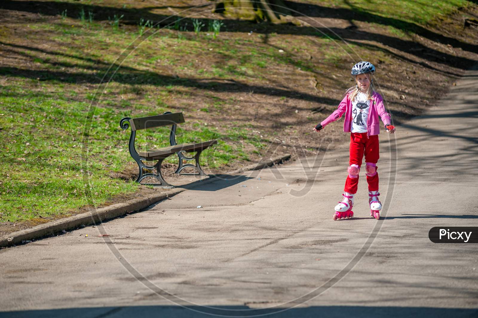 A Young Girl On Roller Blades Passes A Park Bench