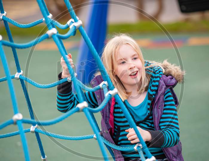 A Happy Young Blonde Girl On A Blue Rope Ladder In A Playground