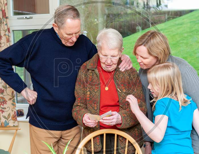 Three Generations Of A Family Gather Around While An Elderly Lady Reads A Birthday Card