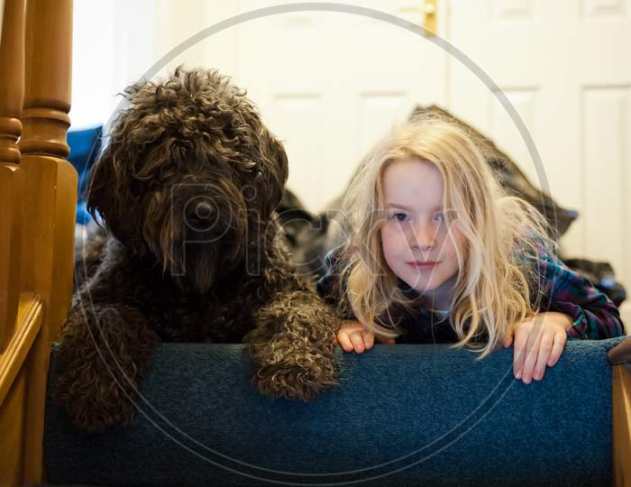 A Young Blonde Girl And A Shaggy Black Labradoodle Looking Down From The Top Of The Stairs
