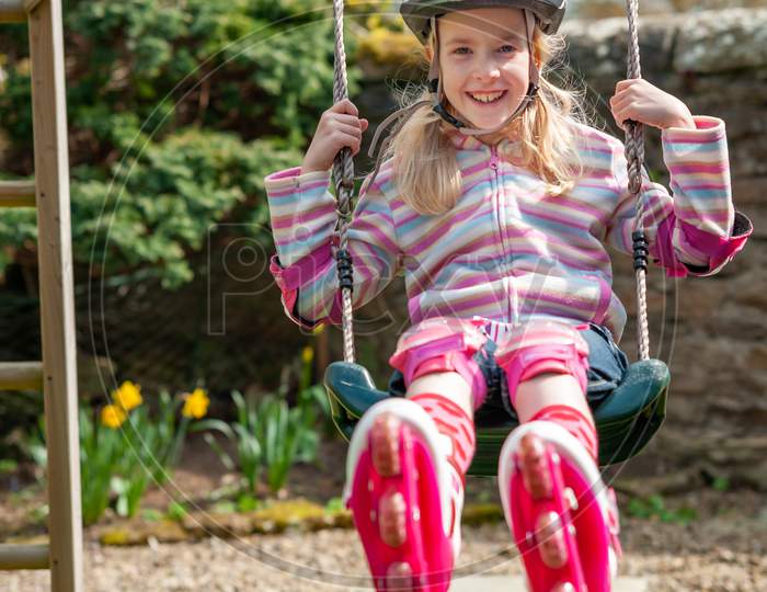 A Smiling Young Blonde Girl Wearing Roller Blades And Helmet While Swinging On A Garden Swing