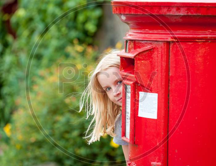 A Young Blonde Girl Peeks Around A Traditional Red British Post Box