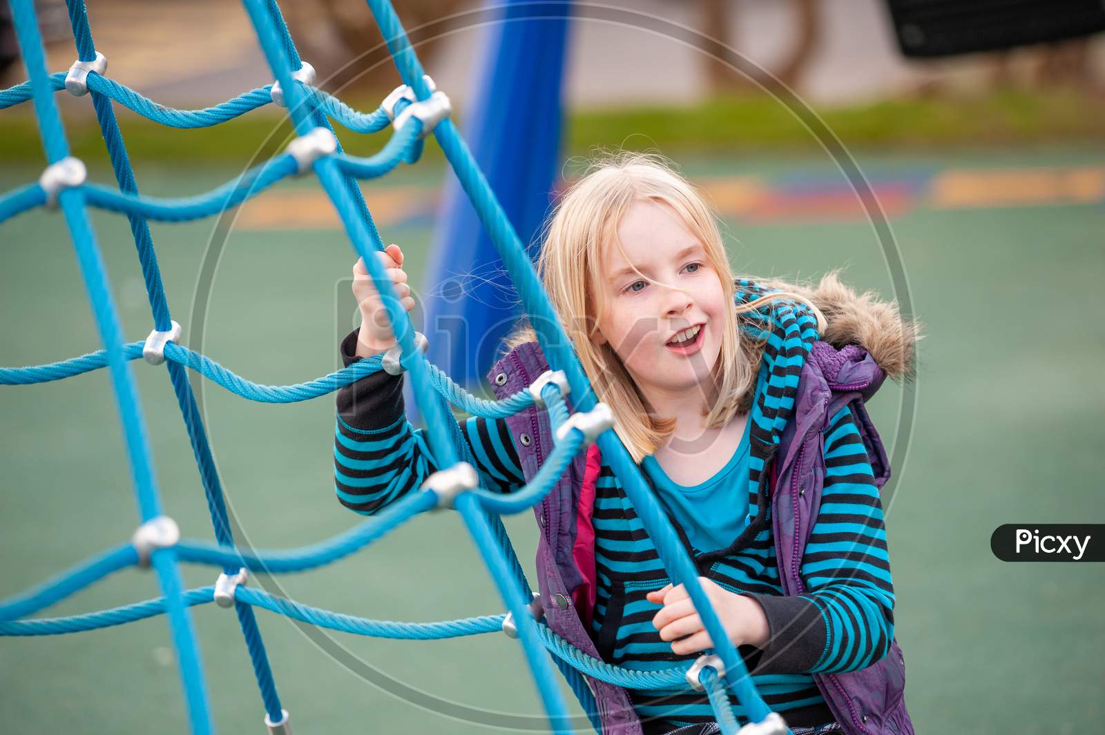 A Happy Young Blonde Girl On A Blue Rope Ladder In A Playground