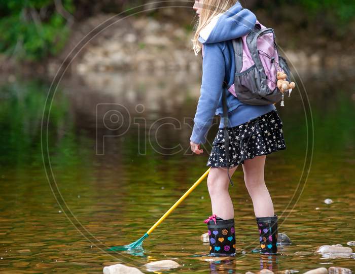 A Young Blonde Girl With Backpack And Fishing Net Wades In The Shallow Water Of A River