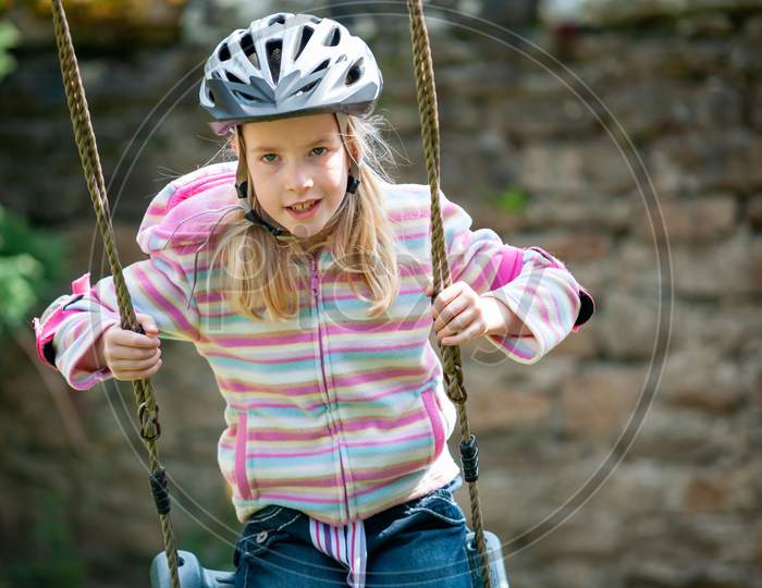 A Young Blonde Girl Wearing A Cycle Helmet While Swinging On A Garden Swing