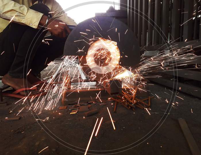 Worker Cutting Iron Rod With Circular Electric Saw With Flying Sparkles In Manufacturing Workshop