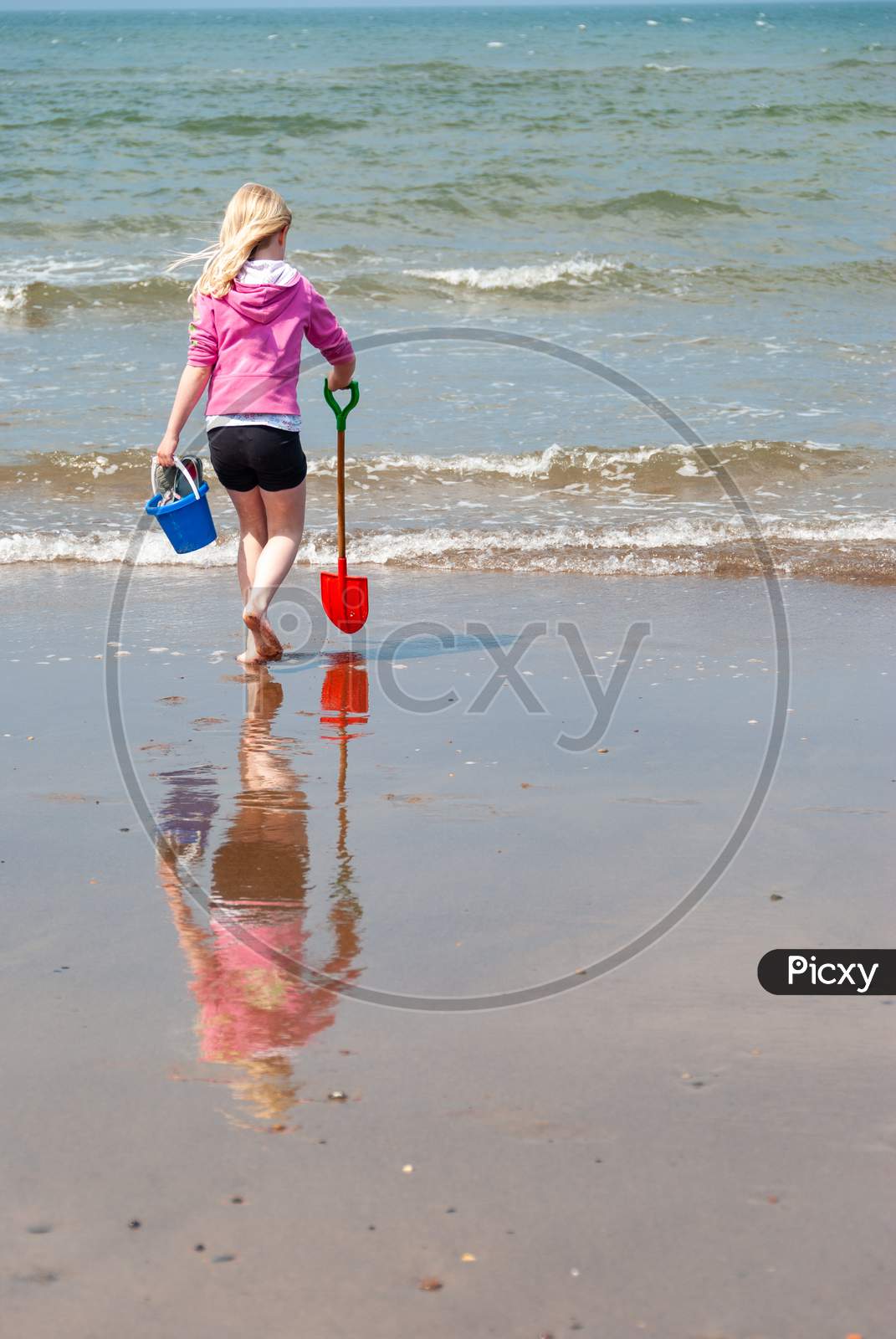 A Young Girl Carries A Bucket And Spade To The Shoreline On A Beach