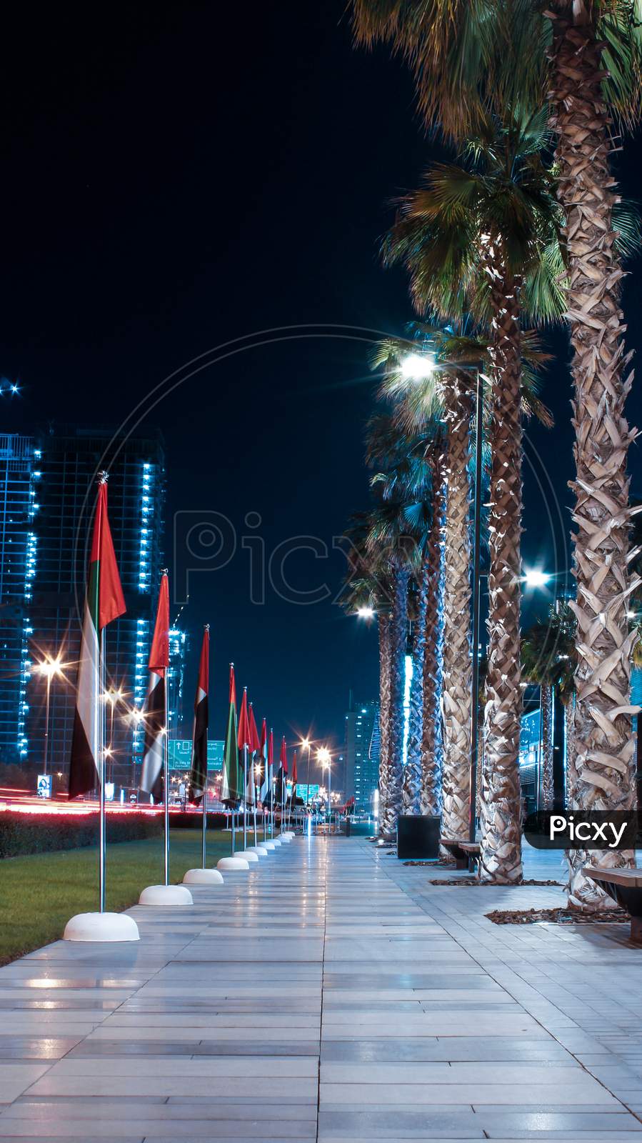 A Symmetrical Long Exposure Photo Of UAE Flags And Palm Trees Alongside A Road And Garden In Downtown Dubai, United Arab Emirates.