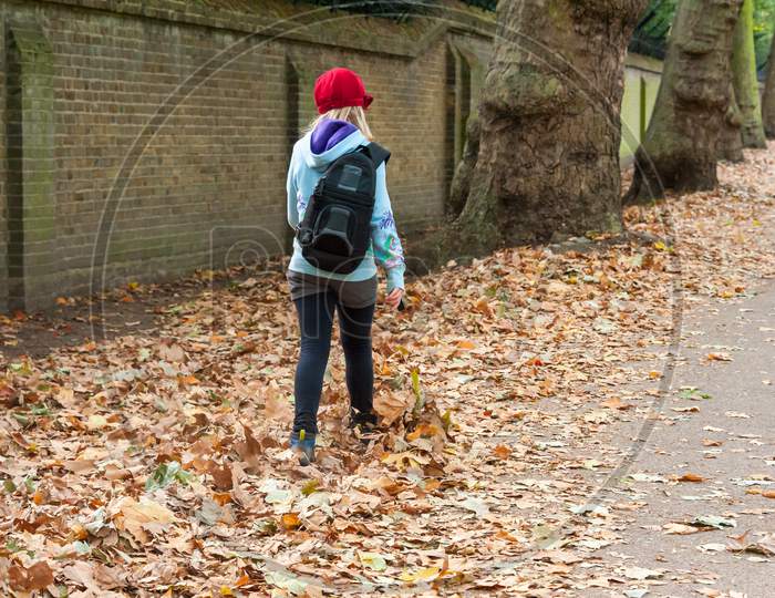 A Young Girl With Backpack Kicks Autumn Leaves While Walking Along A Tree Lined Path