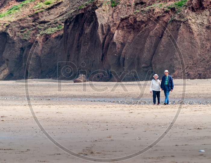 An Elderly Couple Walk Across An Empty Beach At The Base Of Tall Cliffs While Holding Hands