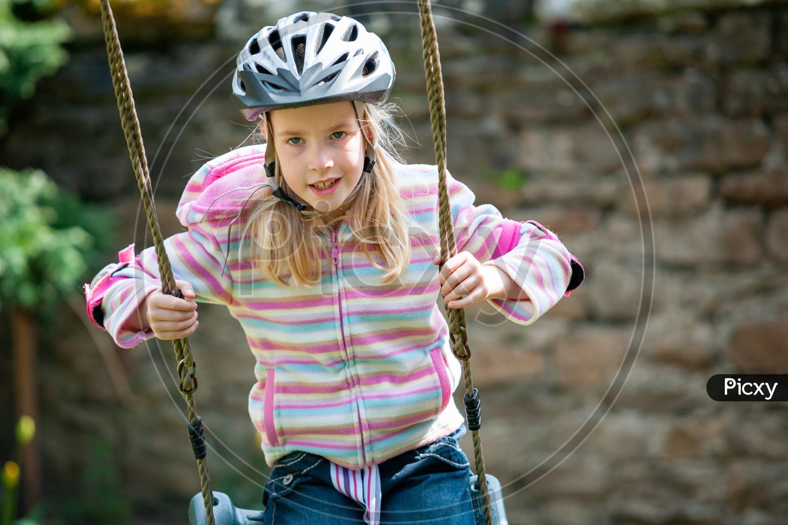 A Young Blonde Girl Wearing A Cycle Helmet While Swinging On A Garden Swing