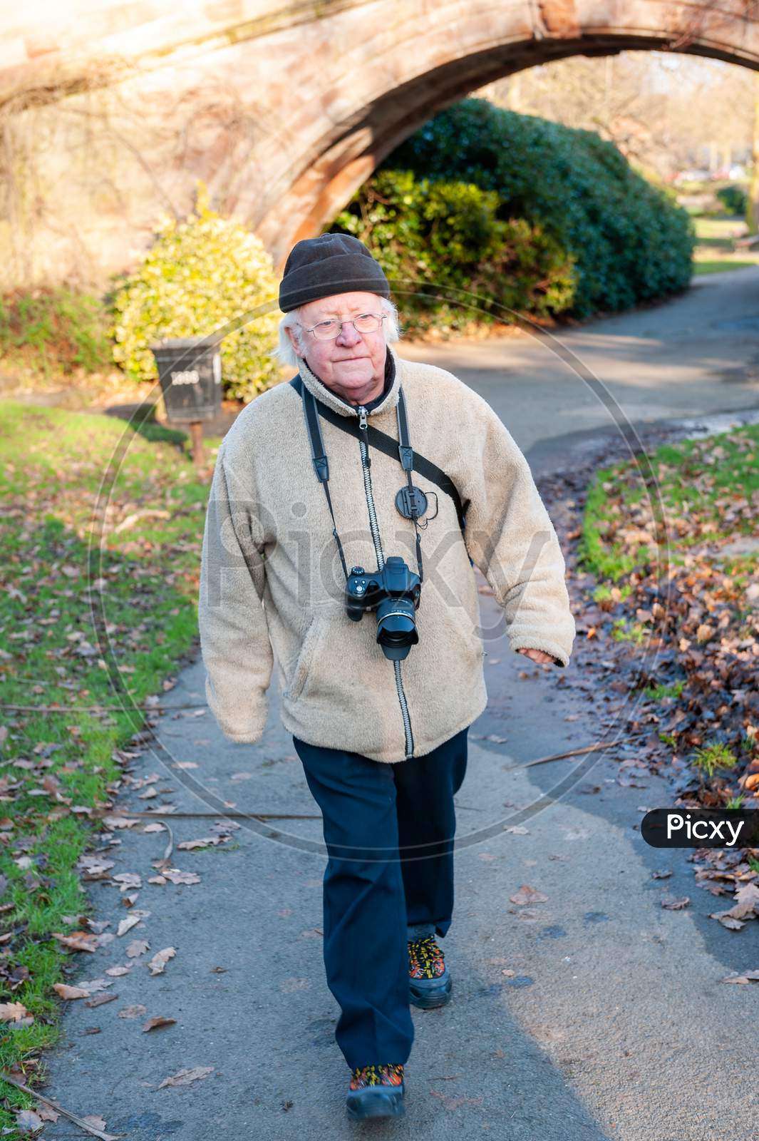 An Elderly Man With A Camera Around His Neck Walks Along A Country Path