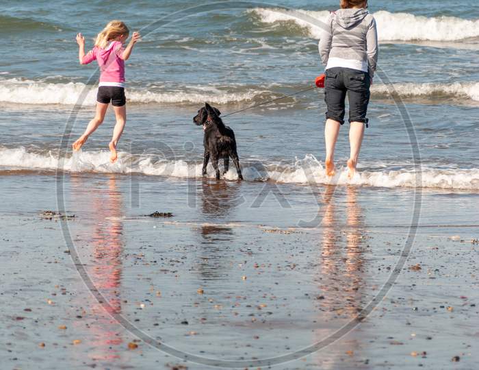 A Mother And Daughter With A Dog Jump Over The Breaking Waves On A Beach