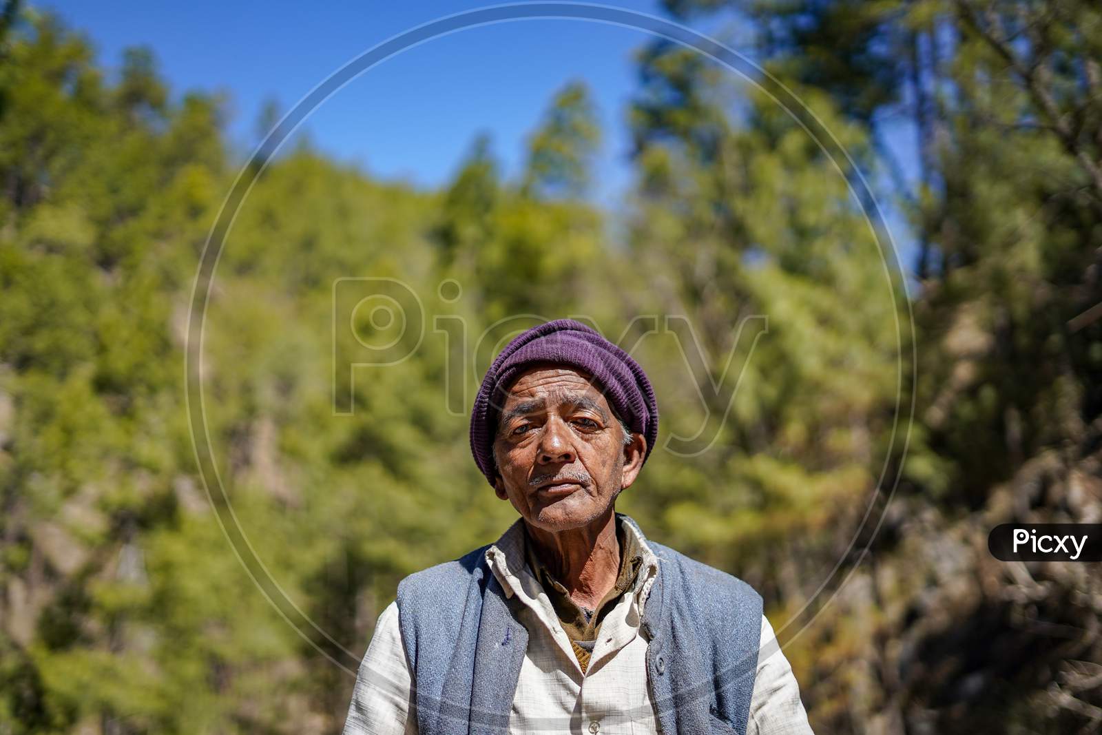 Almora, Uttrakhand / India- June 4 2020: A Wide Portrait Of An Old Man With Wrinkles On His Face Sitting In A Forest Wearing A Cap And A Waistcoat.