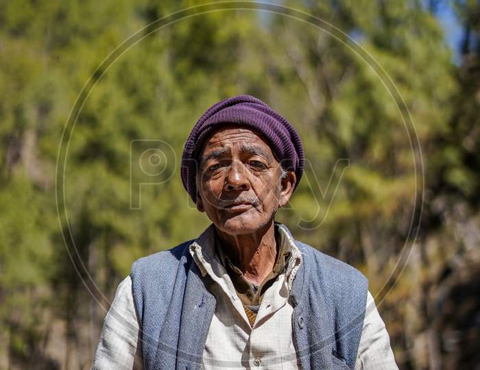 Almora, Uttrakhand / India- June 4 2020: A Wide Portrait Of An Old Man With Wrinkles On His Face Sitting In A Forest Wearing A Cap And A Waistcoat.