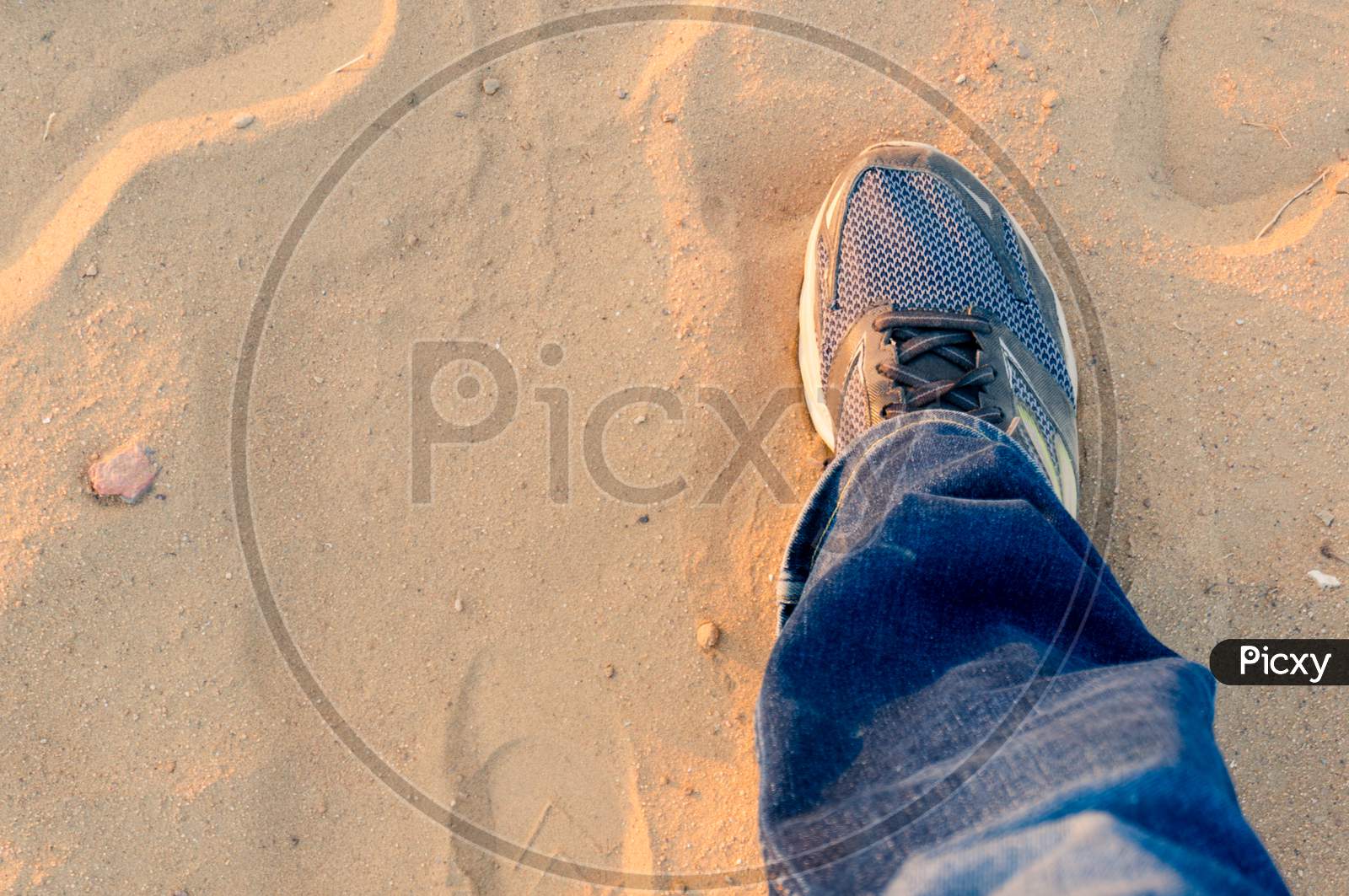Top Down View Of Man Extending A Foot With A Worn Sneaker Jeans On Dusty Sand Showing Travel And Trekking During Evening As Sunlight Bounces Off It