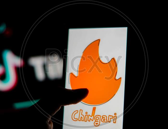 Chingari App Logo On Mobile Screen With Banned Tiktok Application Logo In The Background And A Finger About To Touch