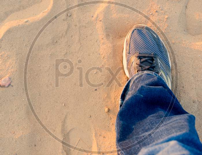 Top Down View Of Man Extending A Foot With A Worn Sneaker Jeans On Dusty Sand Showing Travel And Trekking During Evening As Sunlight Bounces Off It
