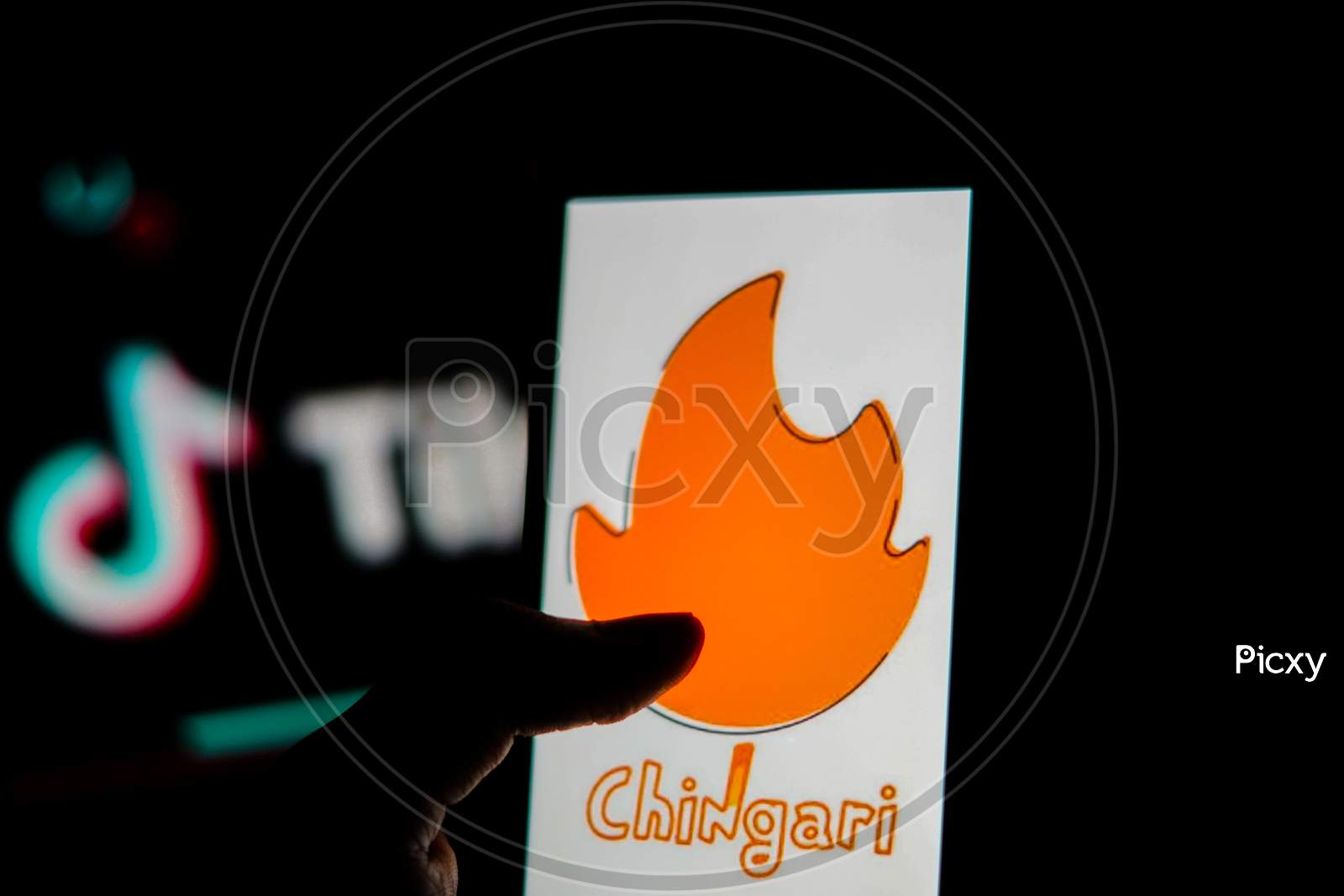 Chingari App Logo On Mobile Screen With Banned Tiktok Application Logo In The Background And A Finger About To Touch