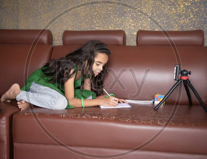 Concept Of Improper, Lazy And Way Of Studying During Homeschooling Or E-Learning And Make Kids Sleepy Or Boredom, Young Girl Busy In Writing By Looking Into Mobile On Sofa During Covid-19 Pandemic