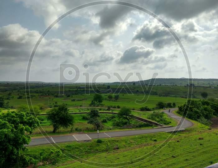 Landscape View Of An Agriculture Field In A Hilly Rural Area. A Narrow Asphalt Road Has Gone Through Its Side.