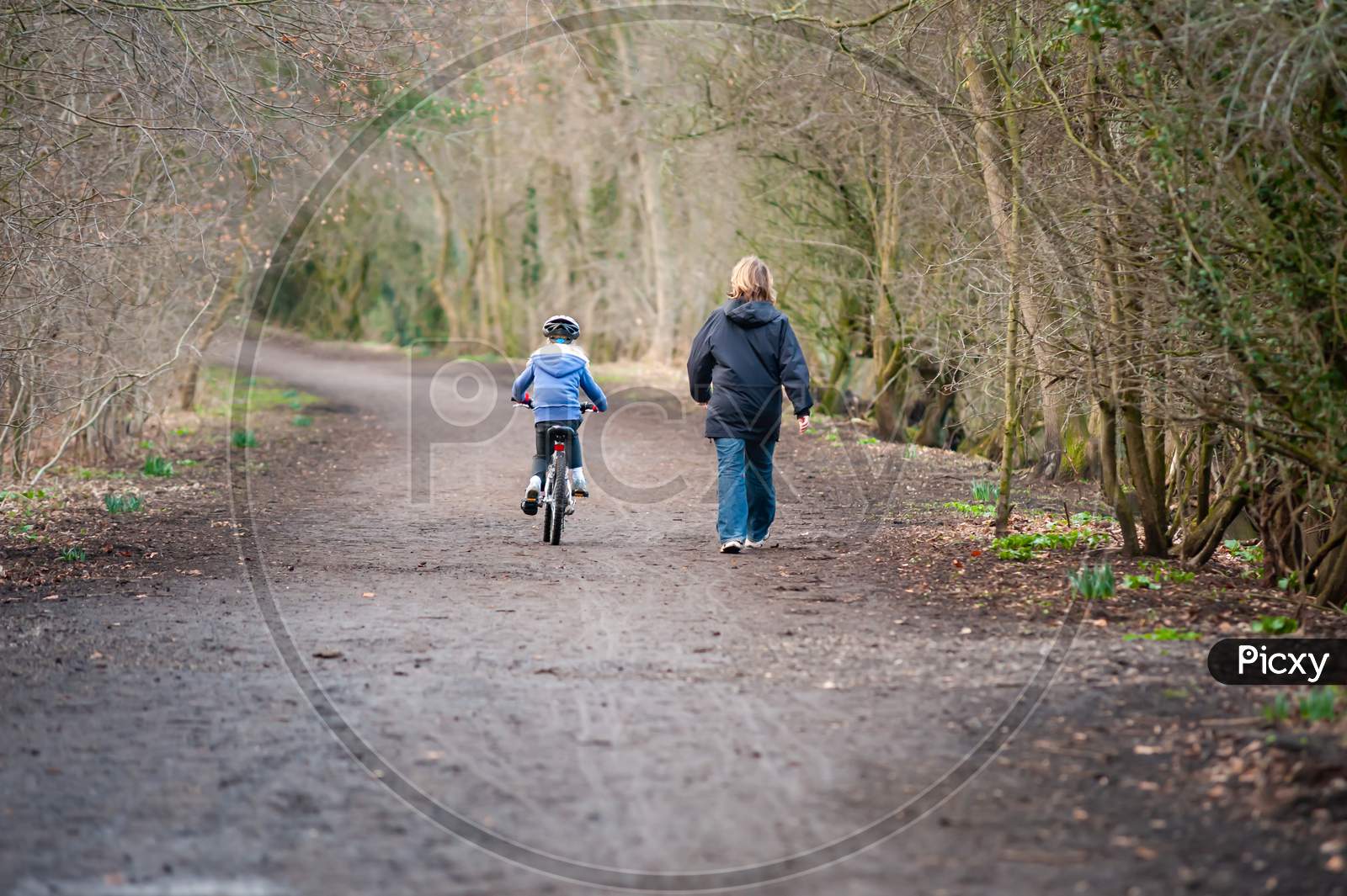 Young Girl Riding Bike Next To Mum Along Country Path