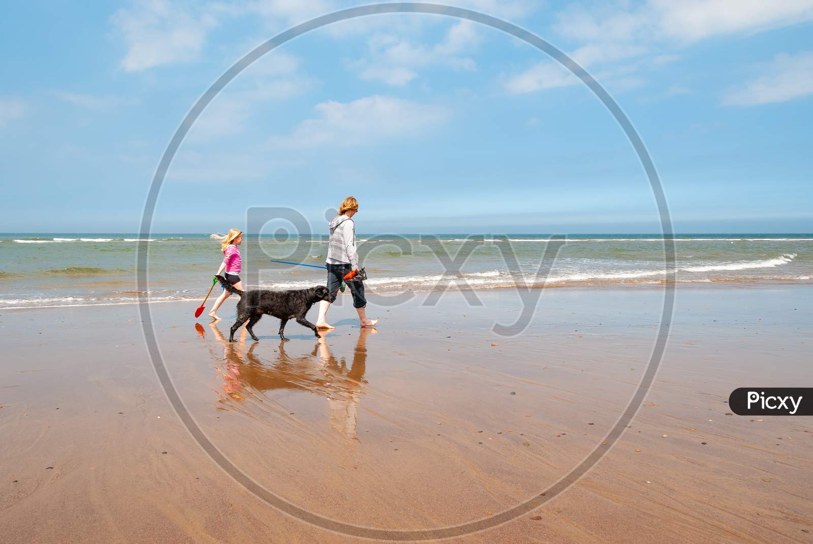 Mother And Daughter Walking Dog Along Sandy Beach Shoreline