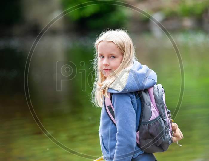 Pretty Young Blonde Girl Looking Back Over Shoulder With Backpack On. Out Of Focus River Scene In Background