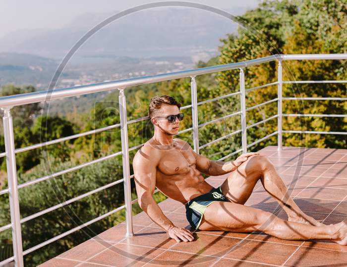 Fashion Portrait Of A Muscular Man Posing Shirtless In Swim Trunks And Sunglasses